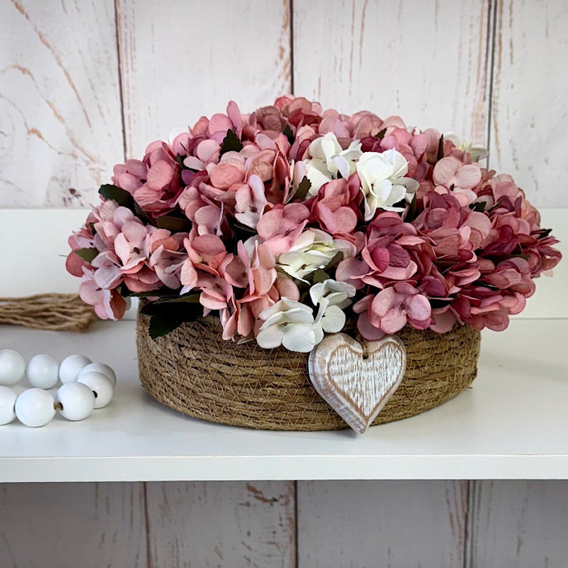 CESTO REDONDO M EM PALHA COM HORTÊNSIAS ENVELHECIDAS ROSADAS E CORAÇÃO 16AX32L/CM